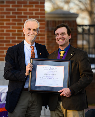 Provost Jones and Dr. Powell holding the Award for Excellence in front of a brick column outside.   