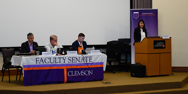 President of the Faculty Senate stands at a podium next to a table with Faculty Senate tablecloth where 3 men are sitting