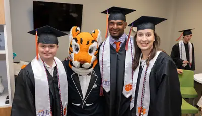 group of Clemson Life students in regalia with their arms around the Tiger mascot