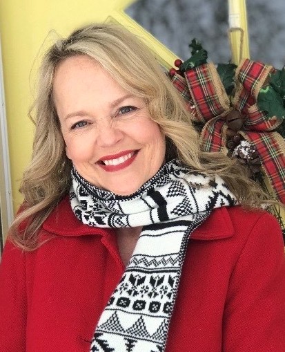 A woman with blonde hair smiles warmly while wearing a red coat and a black and white patterned scarf, surrounded by festive decorations.