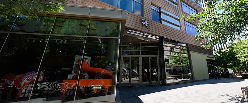 The front of an industrial building with an orange piano seen through large glass windows.