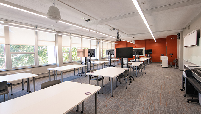 A brightly lit classroom in Lowry Hall featuring multiple, mobile, large monitors; a projector; and room microphones.