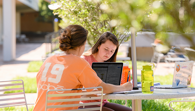 Two students doing schoolwork on laptops while sitting at a small, round outdoor table surrounded by greenery.