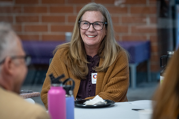 A smiling woman with long, wavy hair and glasses wearing a brown cardigan sits at a table, engaging in conversation during an event.