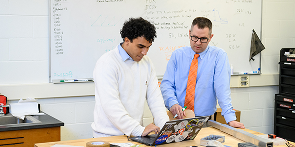 Professor and student collaborating over a laptop in a classroom setting, discussing project details noted on a whiteboard in the background.