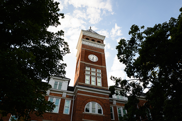 Historic Tillman clock tower of a red brick building surrounded by lush green trees under a blue sky.