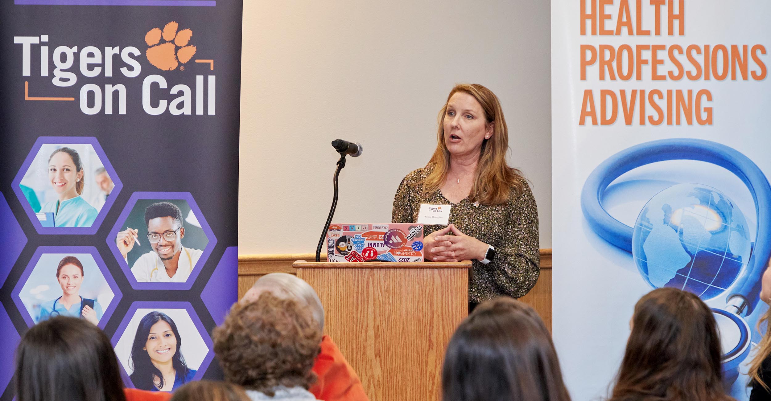 A woman making a presentation to an audience with two banners, Tigers on Call and Health Professions Advising, flanking her.