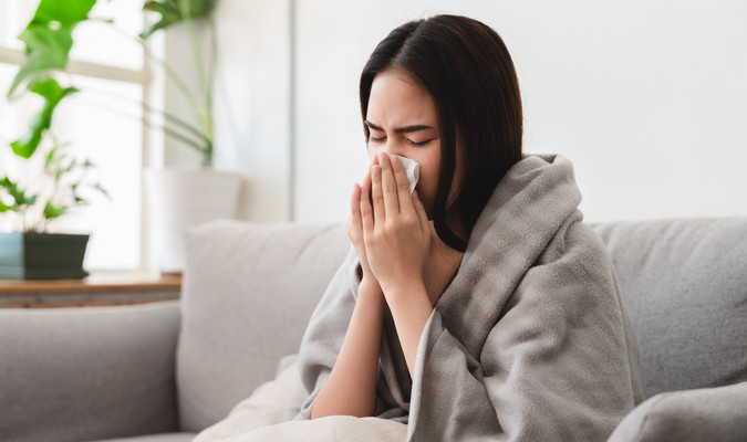 sick asian young woman sneezing into tissue paper while covered with a blanket and sitting on sofa in the living room