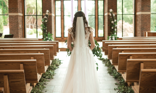 Bride walking down aisle covered with greenery and white flowers.