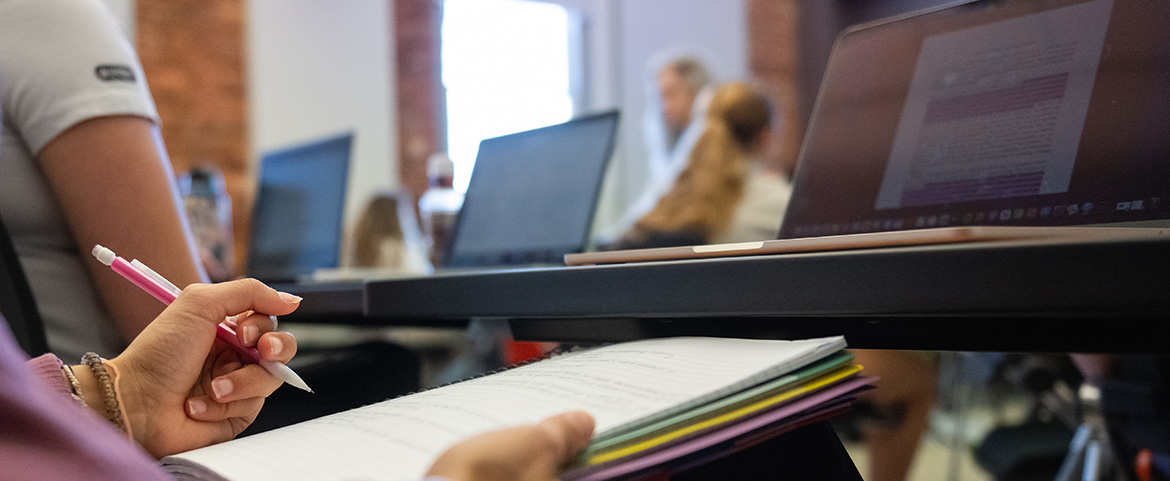 Student in a classroom holding a notebook on her lap and a laptop sitting on the table in front of her