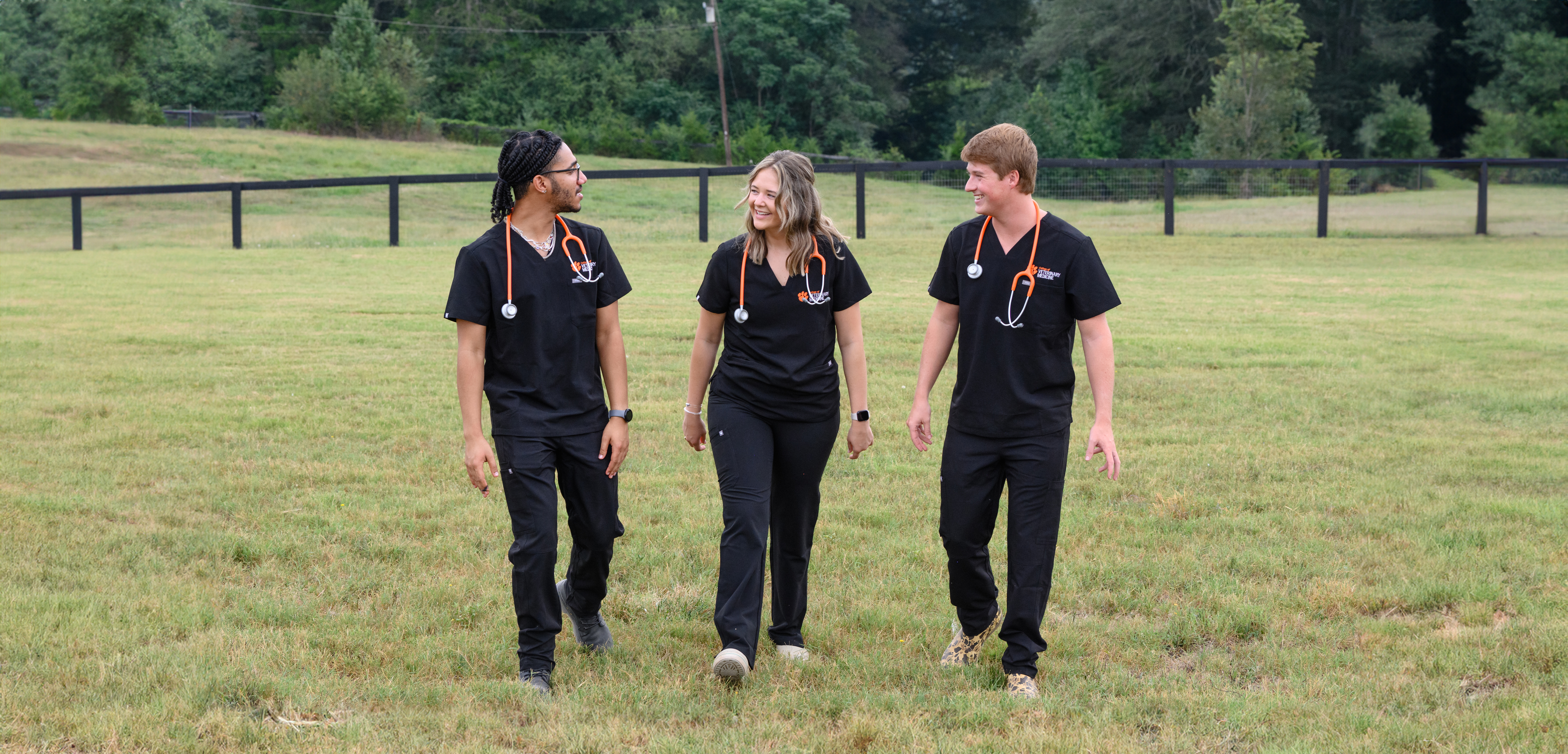 Three students in scrubs walking together in a field at the Clemson equine farm.