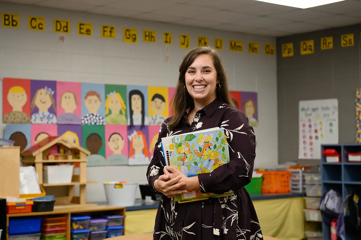A female professor in a dark purple dress with a white floral pattern holds a stack of children’s books while in an elementary school classroom. She stands in front a wall adorned with colorful self-portraits of children created with construction paper and yellow posters of the letters of the alphabet written in black.